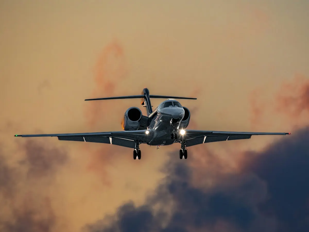 A business jet on final approach with landing lights on against a sunset sky